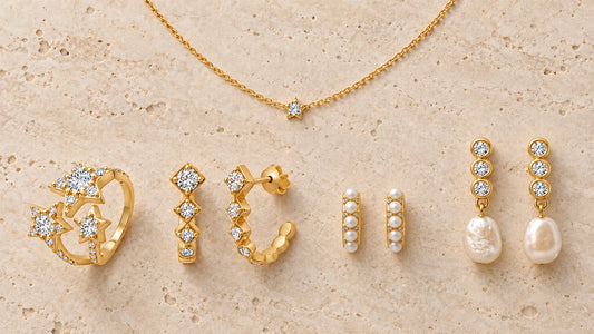 Gold-plated jewelry of different types, hoops, huggies, pearl earrings, an open ring and a necklace, arranged on light travertine stone